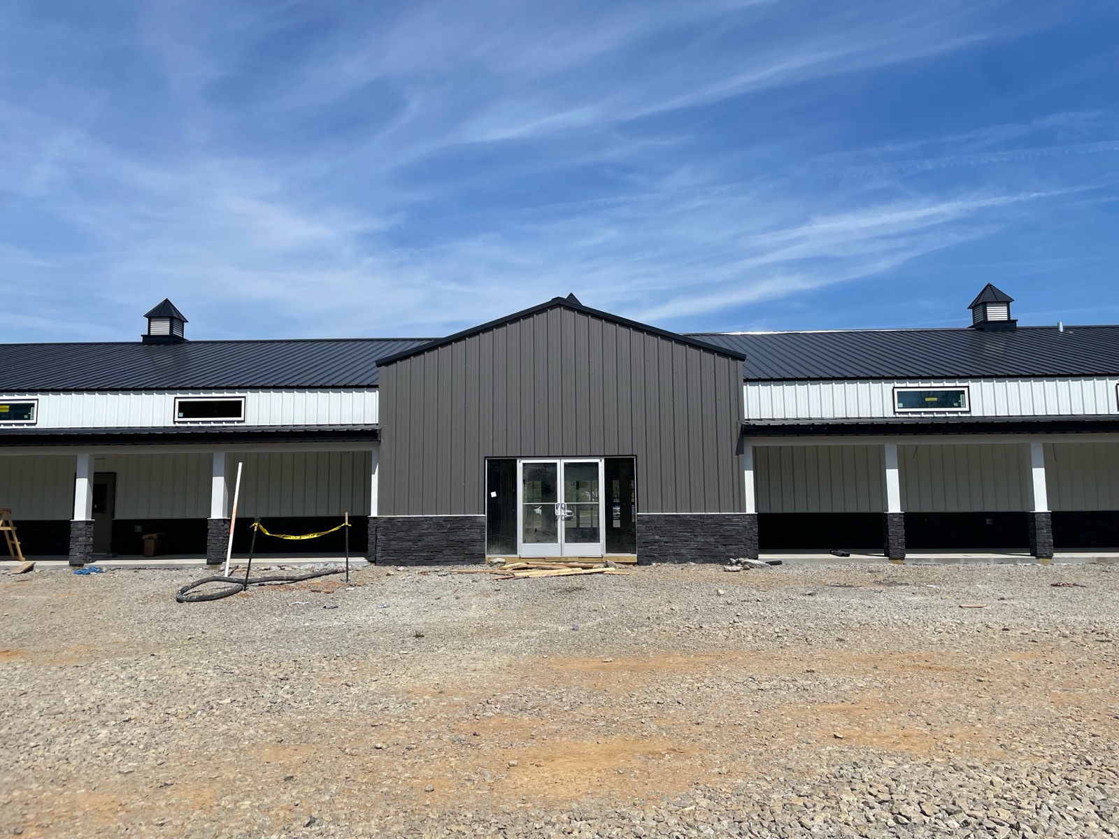 Front view of the main entrance gable at Thompson's Station Hardware, featuring dark gray siding, glass double doors, cupolas on each side, and stone-accented column bases.