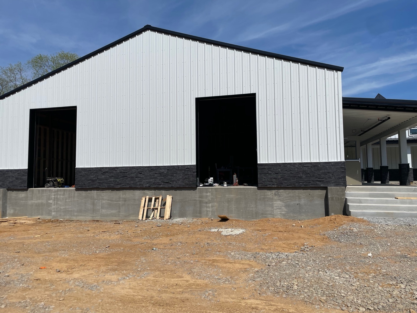Rear exterior gable of Thompson's Station Hardware showing two large open loading bay doorways, concrete foundation walls, and completed white metal siding with a covered porch visible to the right.