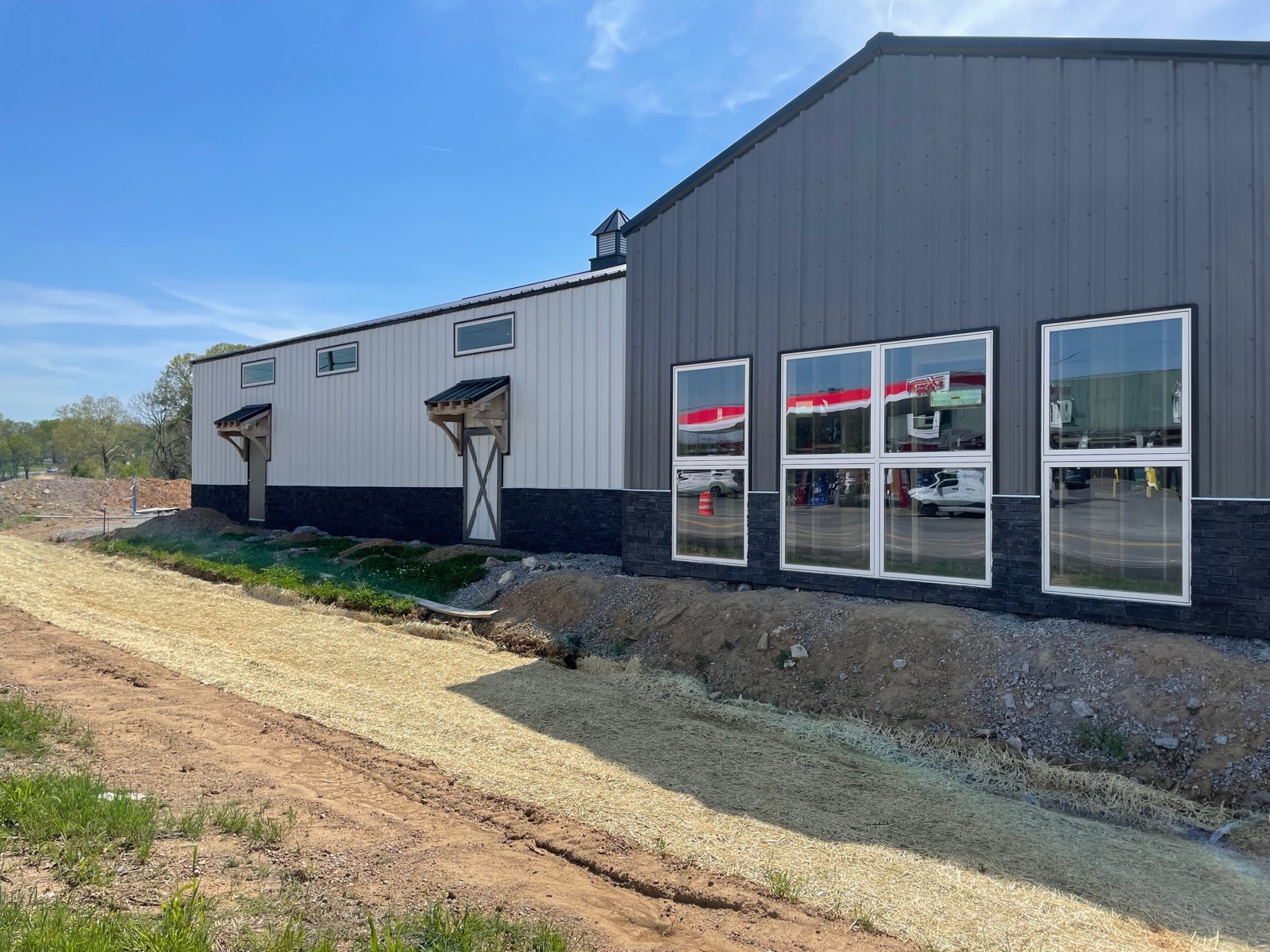 Side exterior of Thompson's Station Hardware showing large showroom windows reflecting the gas station across Lewisburg Pike, with gabled side entries and stone wainscot along the base.