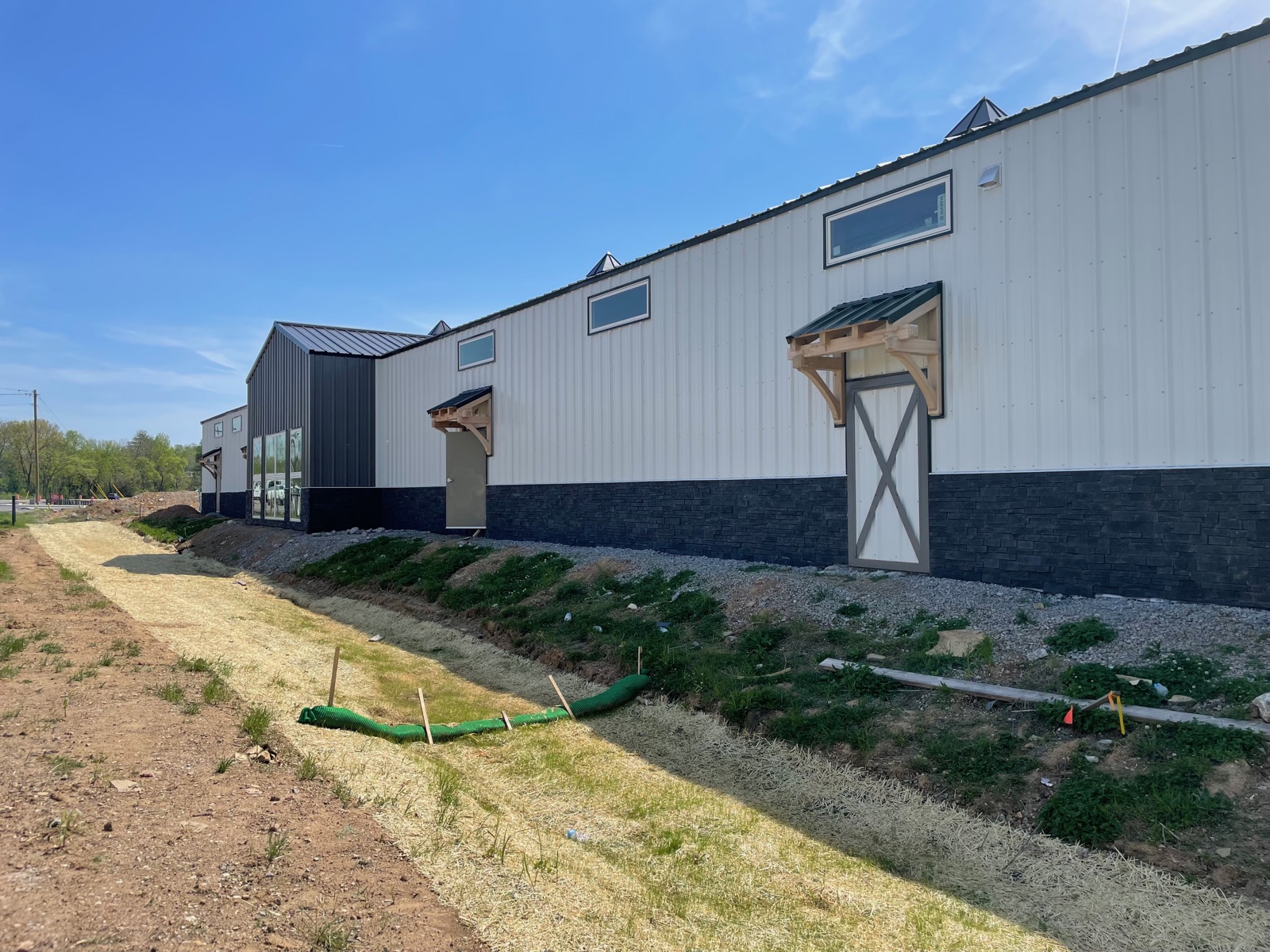 Roadside profile of Thompson's Station Hardware facing Lewisburg Pike, showing gabled accents with timber brackets, stone wainscot, and fresh site grading with erosion control silt fence below.