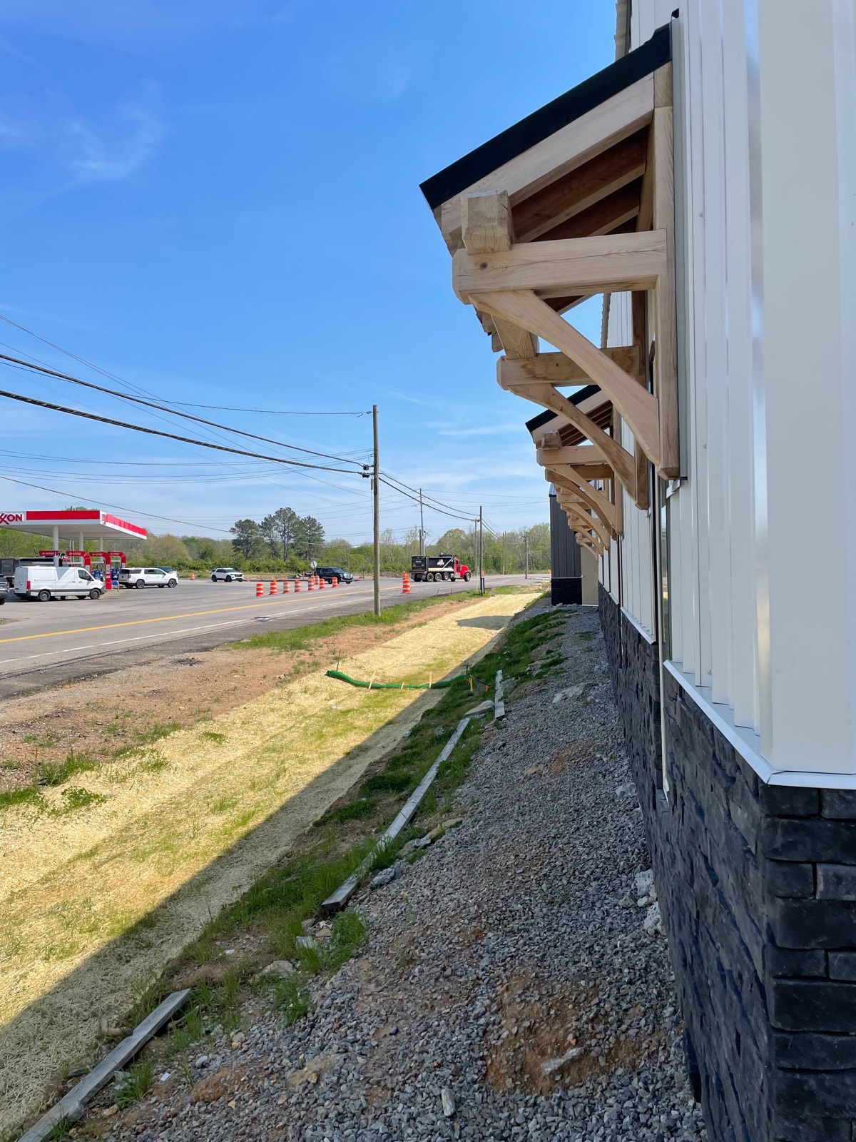 Close-up looking down the front porch roofline of Thompson's Station Hardware, highlighting heavy timber corbel brackets supporting the porch overhang, with Lewisburg Pike visible in the background.