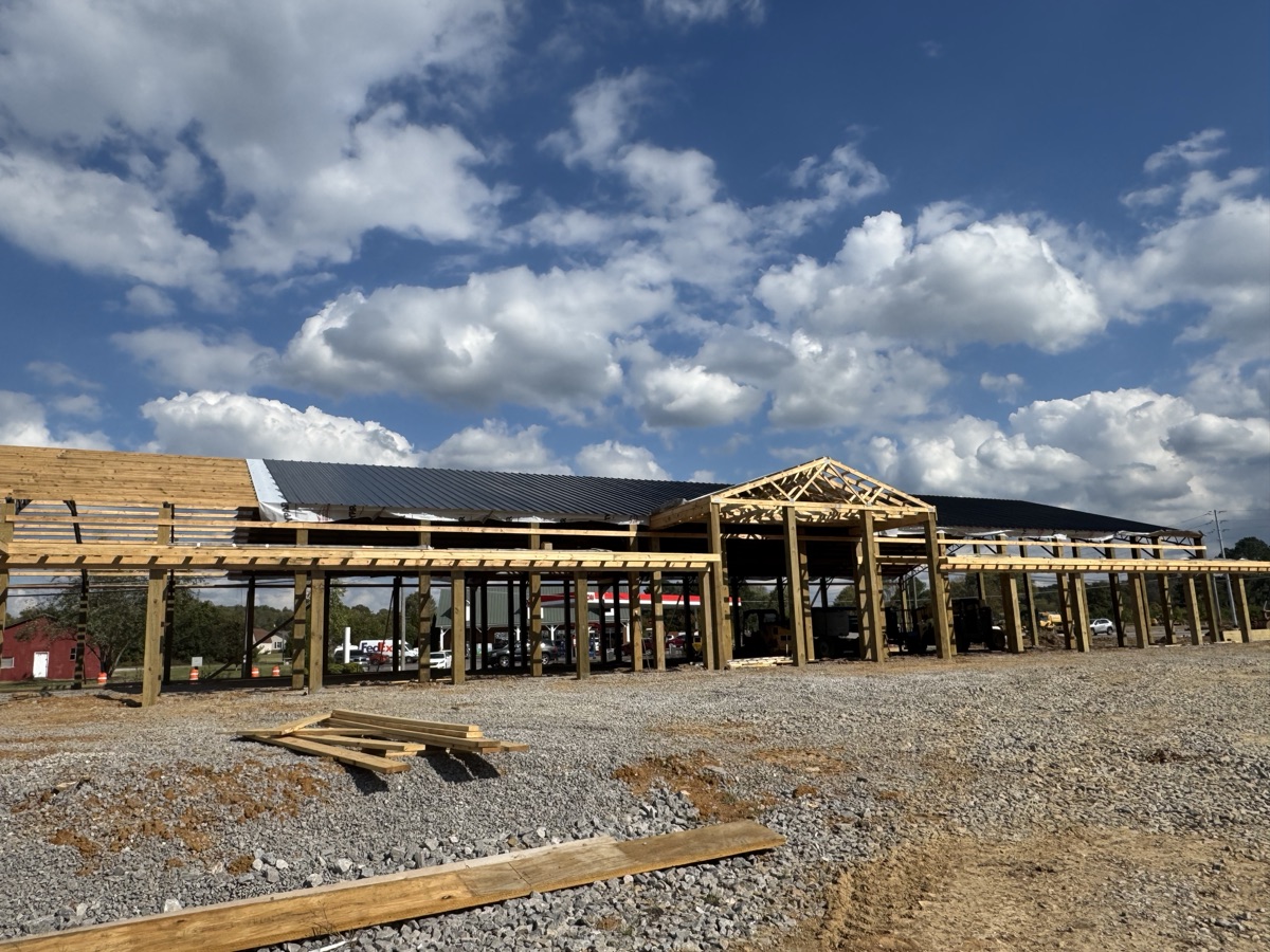 Exterior side view of Thompsons Station Hardware construction in Franklin, TN, showcasing wooden posts and horizontal framing with part of the roof installed under a cloudy blue sky.