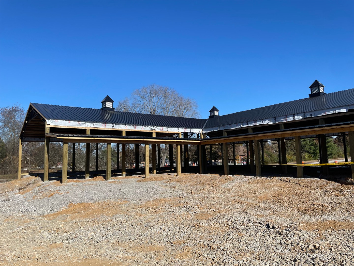 Rear exterior view of Thompson's Station Hardware under construction in Franklin, TN, showing completed metal roof with decorative cupolas and timber post framing against a clear blue sky.