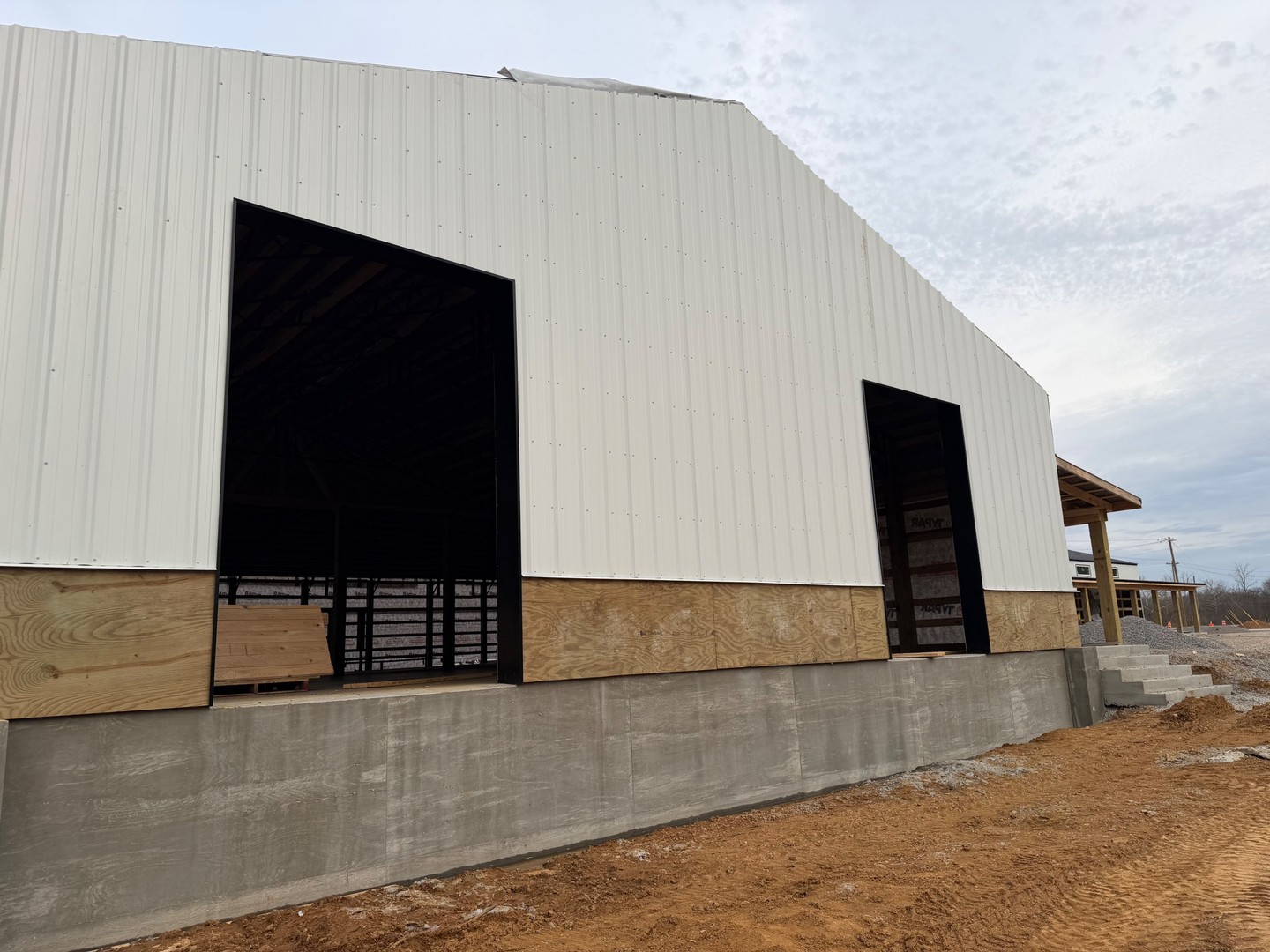 Rear exterior view of Thompson's Station Hardware construction in Franklin, TN, showing loading dock openings with concrete foundation, metal siding, and timber porch framing visible on the right.